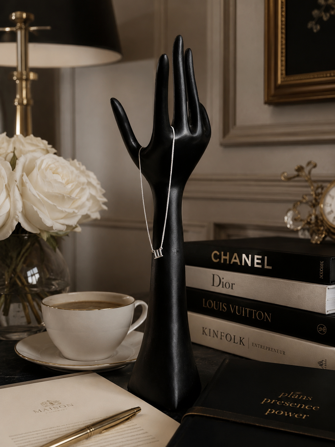 A silver-toned necklace with a small rectangular pendant is draped over a glossy black hand-shaped jewelry display. The display sits on a dark tabletop in a styled interior setting with a white rose bouquet, a cup of coffee, books including Chanel, Dior, Louis Vuitton, and Kinfolk, and a pen on a notebook. 111 Lucky On Purpose Jewelry Jewellery. 