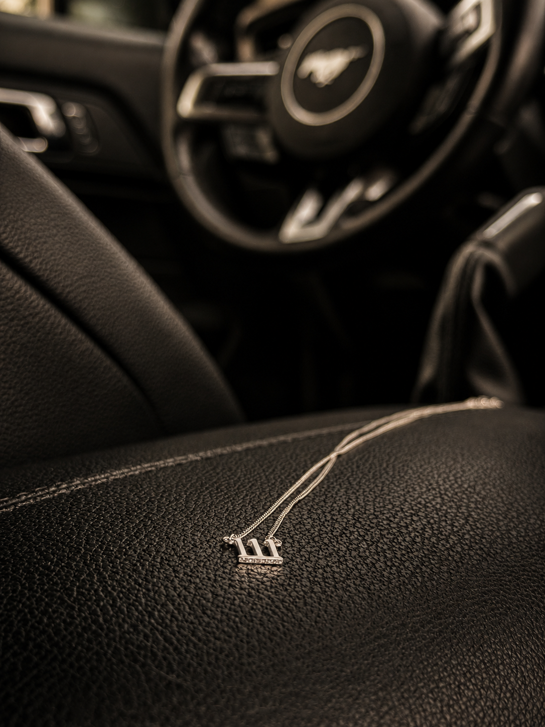 A close-up of a black leather car interior with a small silver metallic emblem on the seat, with a blurred steering wheel featuring a horse logo in the background. 111 Lucky On Purpose Jewelry Jewellery. 