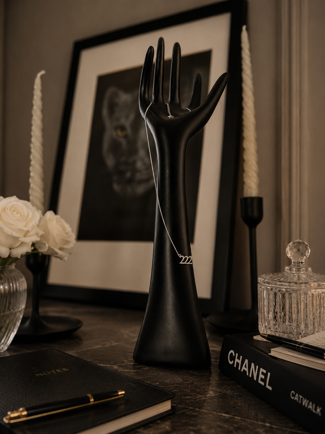 A delicate silver necklace with a triple-zigzag pendant is displayed on a black hand-shaped jewelry stand. The stand is styled on a dark tabletop next to a Chanel book, a glass jar, and candles, with a framed picture in the background. 222 Lucky On Purpose Jewelry Jewellery. 