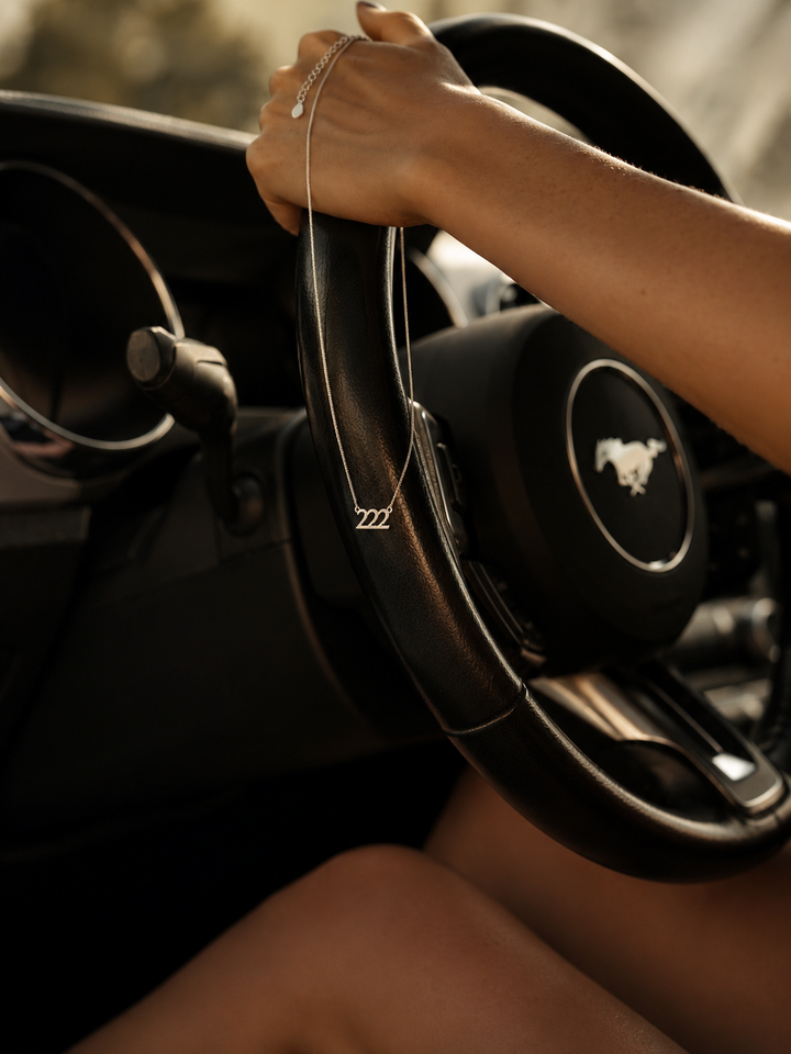 A close-up lifestyle shot of a person's hand resting on a black leather-wrapped steering wheel inside a car. The steering wheel features a black center cap with a silver Mustang horse logo, and the person is wearing a delicate silver chain bracelet. 222 Lucky On Purpose Jewelry Jewellery. 