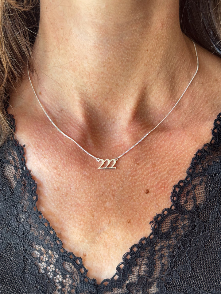 A close-up of a person's neck and upper chest wearing a delicate silver necklace with a small three-stroke initial pendant spelling '222'. The necklace is shown against the skin, with a black lace top visible at the bottom. Lucky On Purpose Jewelry Jewellery. 