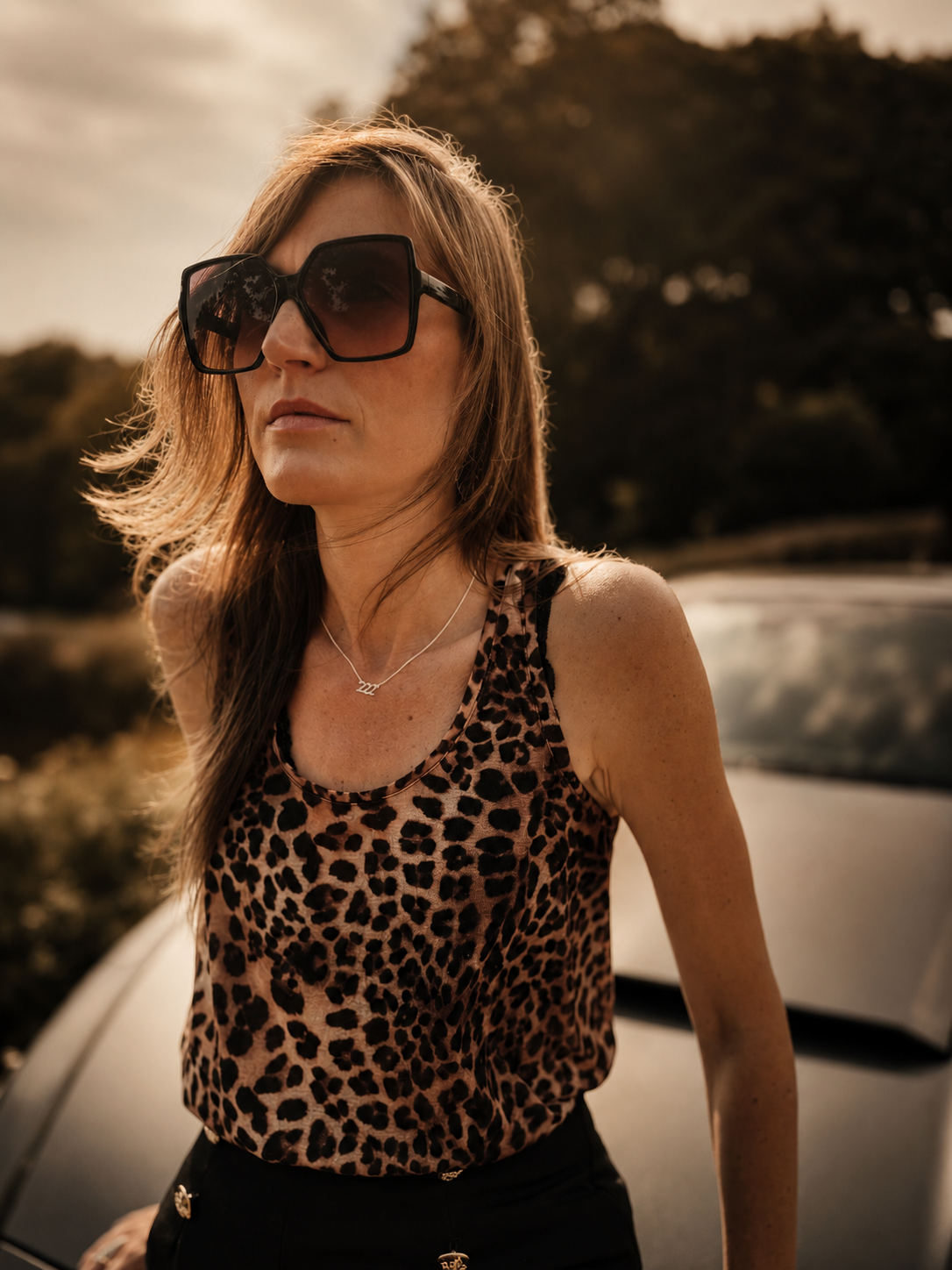 A woman outdoors wearing oversized dark sunglasses and a leopard-print sleeveless top, standing in front of a blurred car and trees. 222 Lucky On Purpose Jewelry Jewellery. 