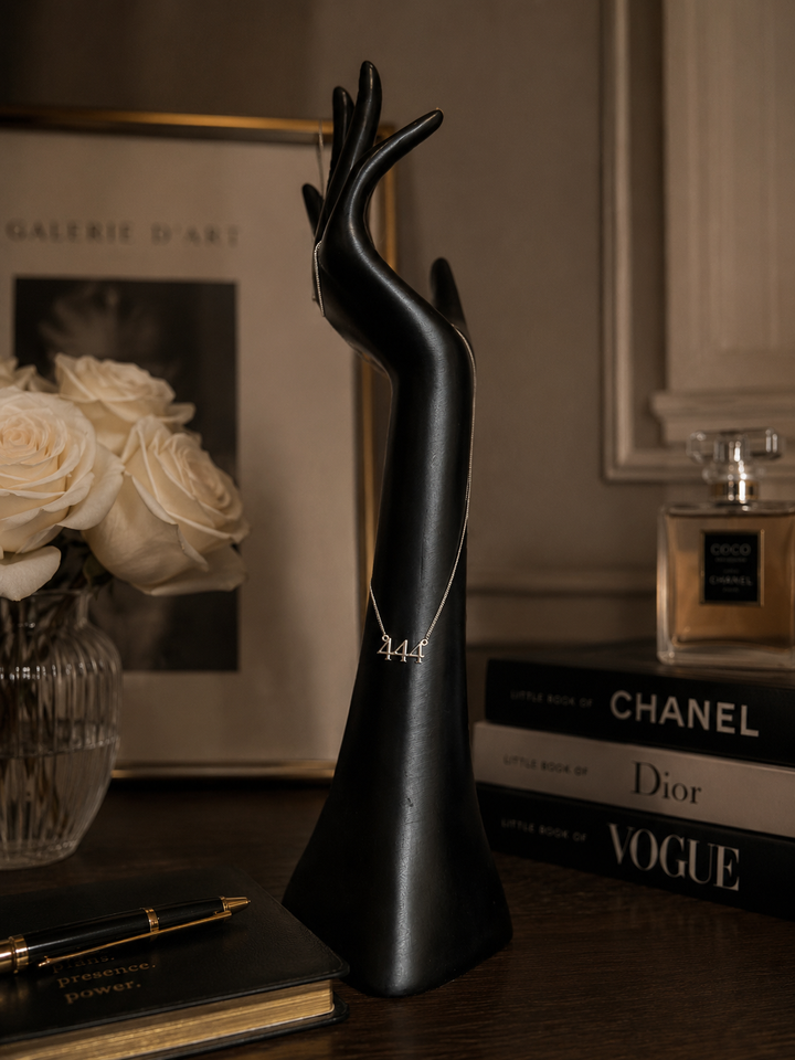 A black sculptural hand-shaped jewelry display stand holding a delicate silver necklace, styled on a dark wooden table with books, a perfume bottle, and white roses in the background. Lucky On Purpose Jewelry Jewellery.  444