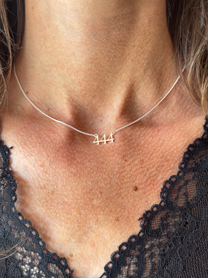 A close-up of a model's neck and chest wearing a delicate silver chain necklace with a small script '444' pendant. Lucky On Purpose Jewelry Jewellery. 
