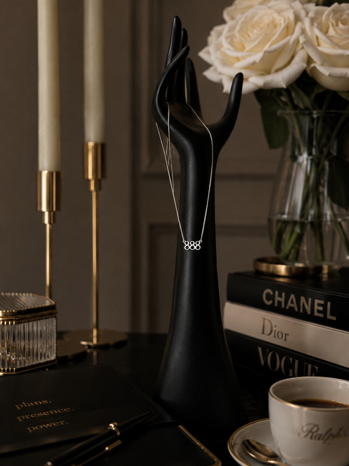 A delicate silver necklace with a small geometric pendant is displayed on a black sculptural hand-shaped jewelry stand. The stand is styled in a sophisticated interior setting with a vase of white roses, gold candlesticks, a Chanel and Dior book stack, and a Ralph Lauren cup on a dark table. Lucky On Purpose Jewelry Jewellery. 888