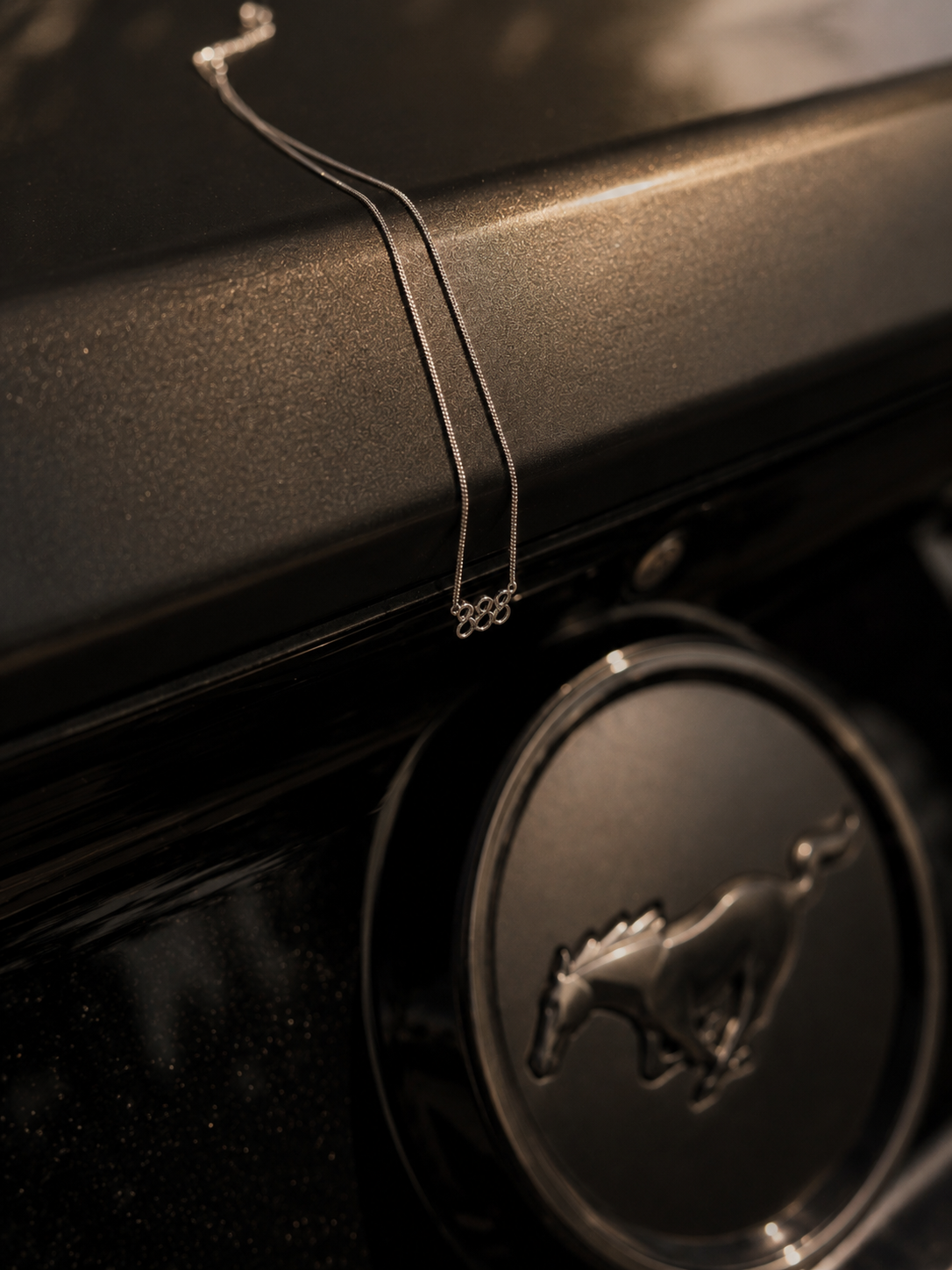 A delicate silver chain necklace with a small three-ring pendant draped over the glossy black hood of a car, positioned near the Mustang emblem. Lucky On Purpose Jewelry Jewellery. 888 