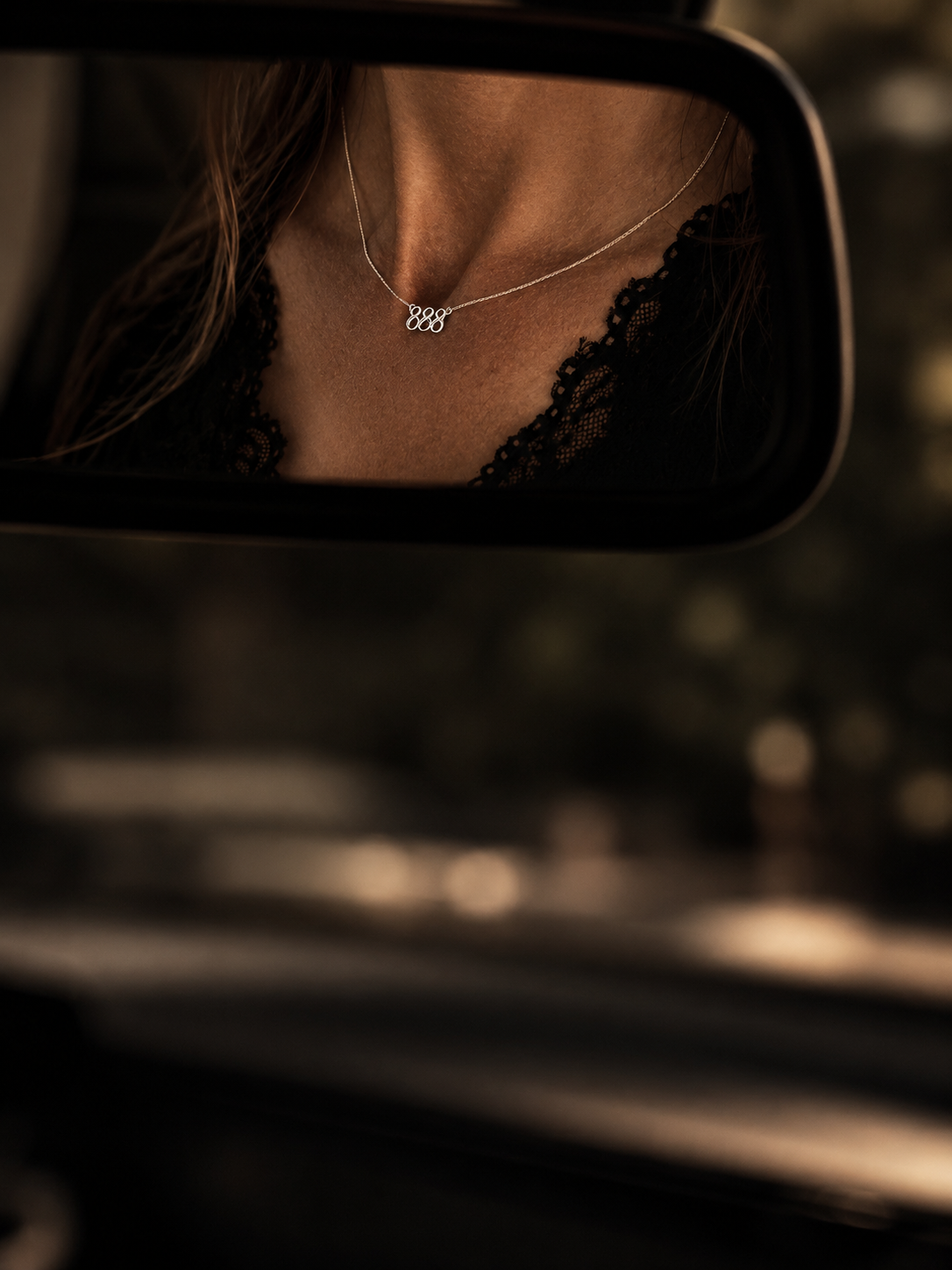 A close-up of a woman's neck and chest wearing a delicate silver necklace with a small pendant, reflected in a car rearview mirror. She is wearing a black lace top, with a blurred car interior and outdoor background. Lucky On Purpose Jewelry Jewellery. 888